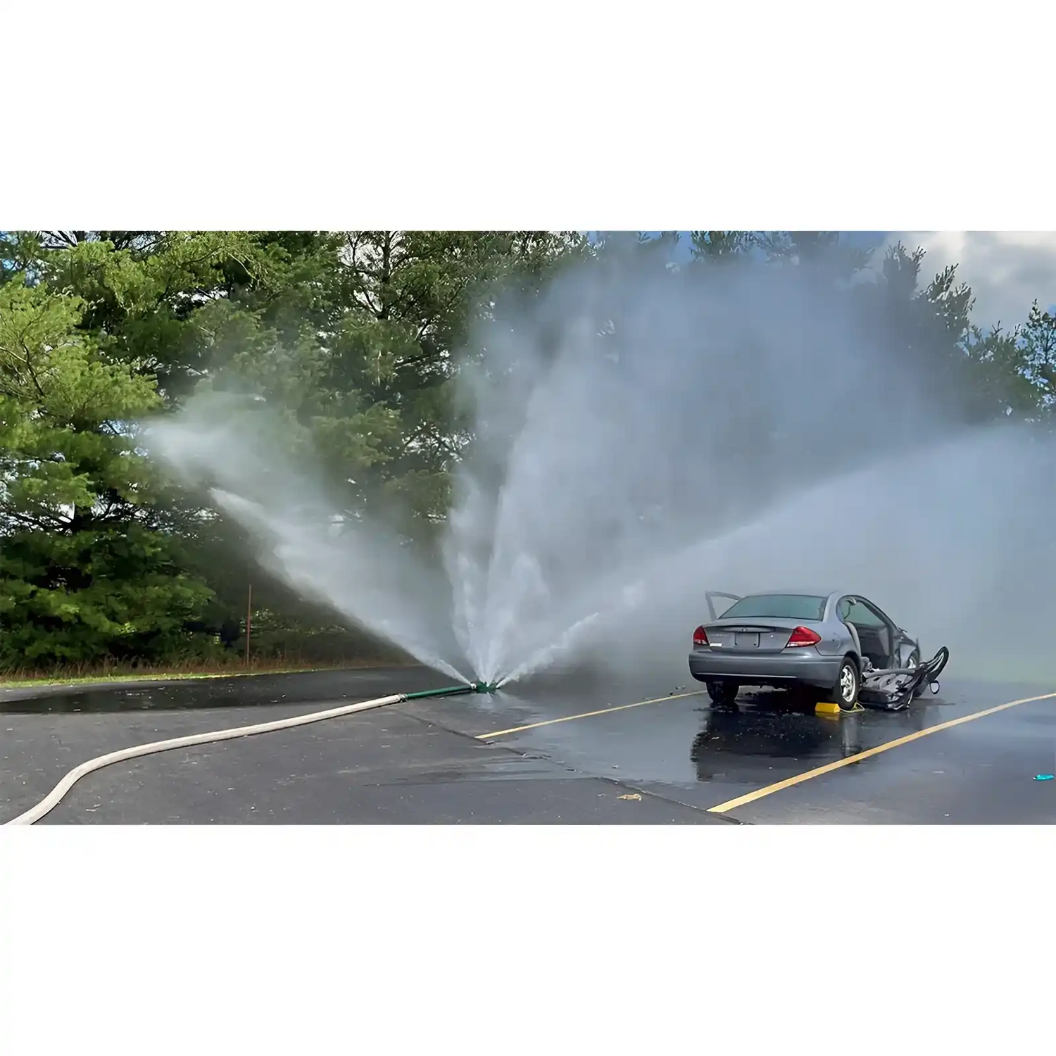 A broken fire hydrant spraying water forcefully across a road near a parked car.