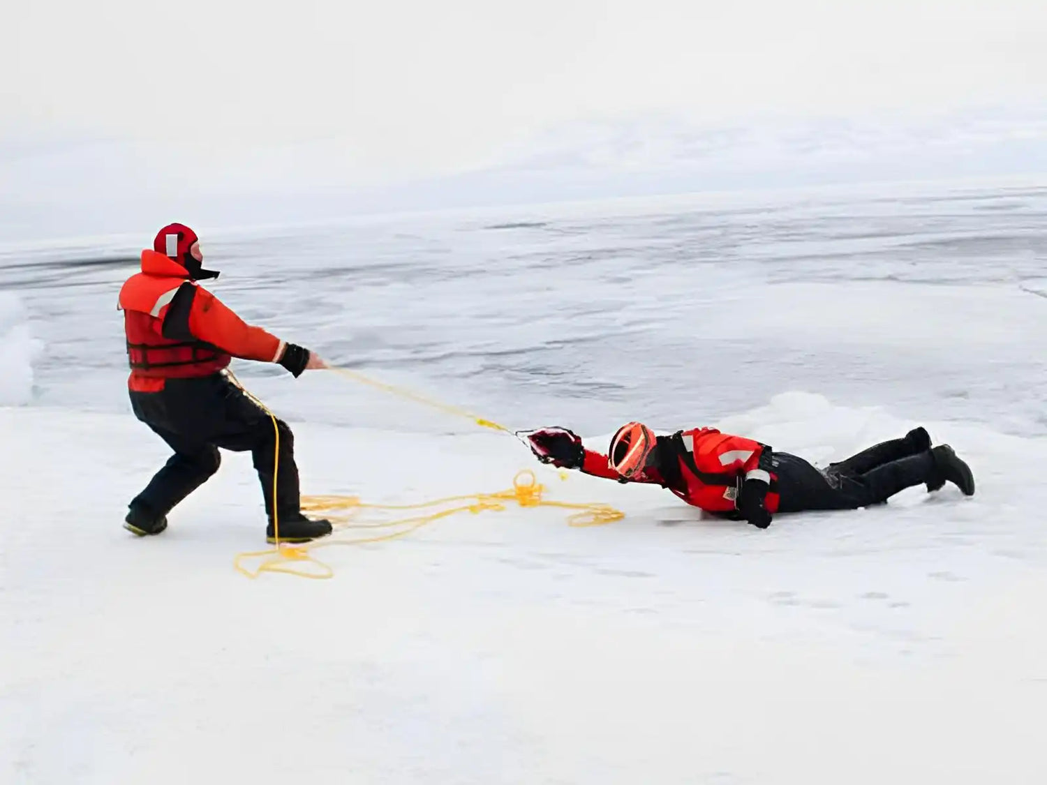 Ice rescue training exercise with one person pulling another across frozen water using safety ropes.