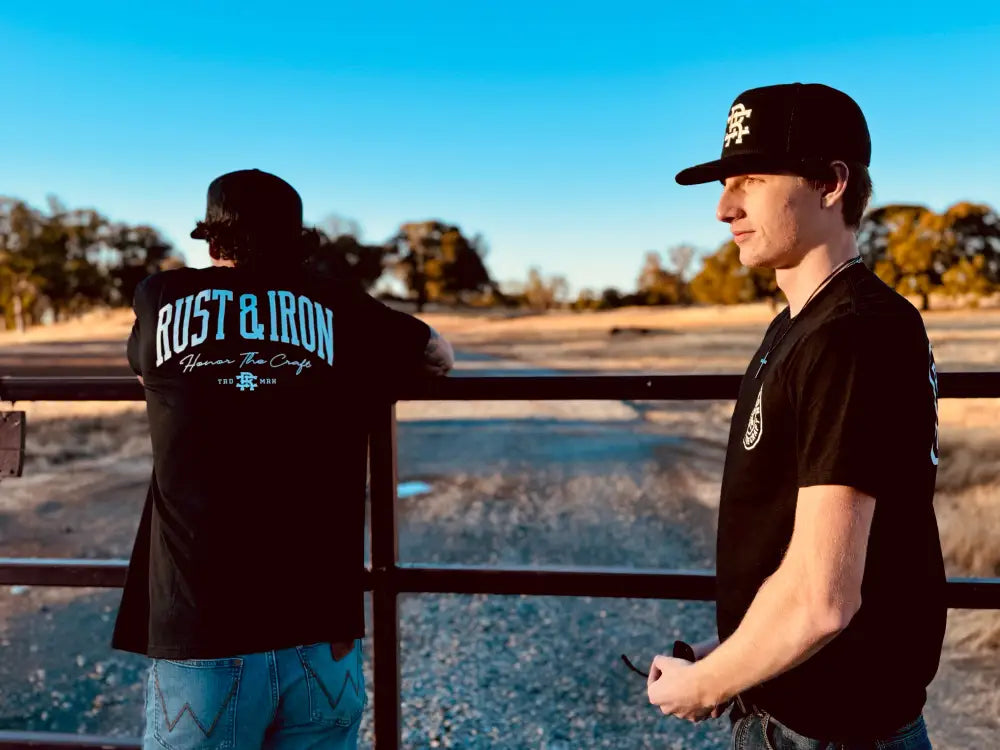 Two people in black staple tees by a railing, one featuring RUST & IRON text