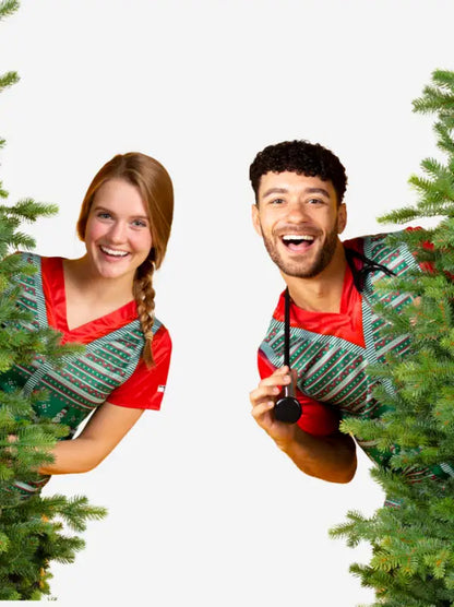 Two people in matching red ugly holiday sweaters peek through evergreen branches